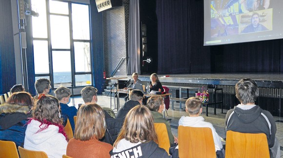 Rund 60 Neuntklässler des JAG haben sich in der Aula eingefunden, um mit dem Autor David A. Robertson (oben rechts) ins Gespräch zu kommen. Er ist via Zoom live von Kanada zugeschaltet. Moderatorin Rikarde Riedesel (rechts) und Bettina Born – sie trägt die deutschen Texte vor. (WP-Foto: Eberhard Demtröder)